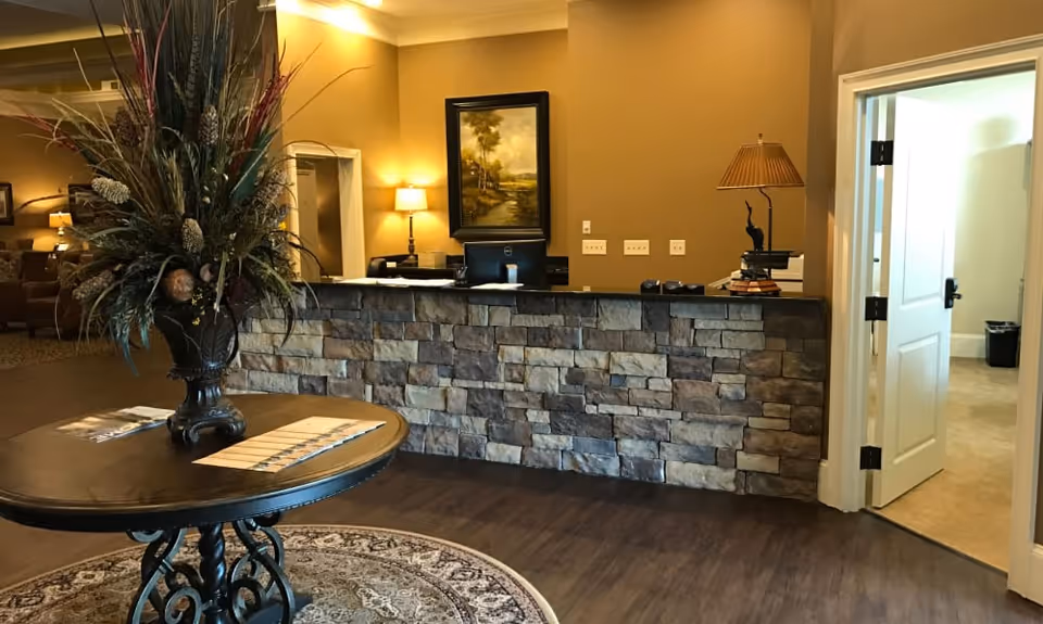 Reception area with a stone front desk, a round wooden table with a large floral arrangement, warm lighting, a painting on the wall, and an open door leading to another room.