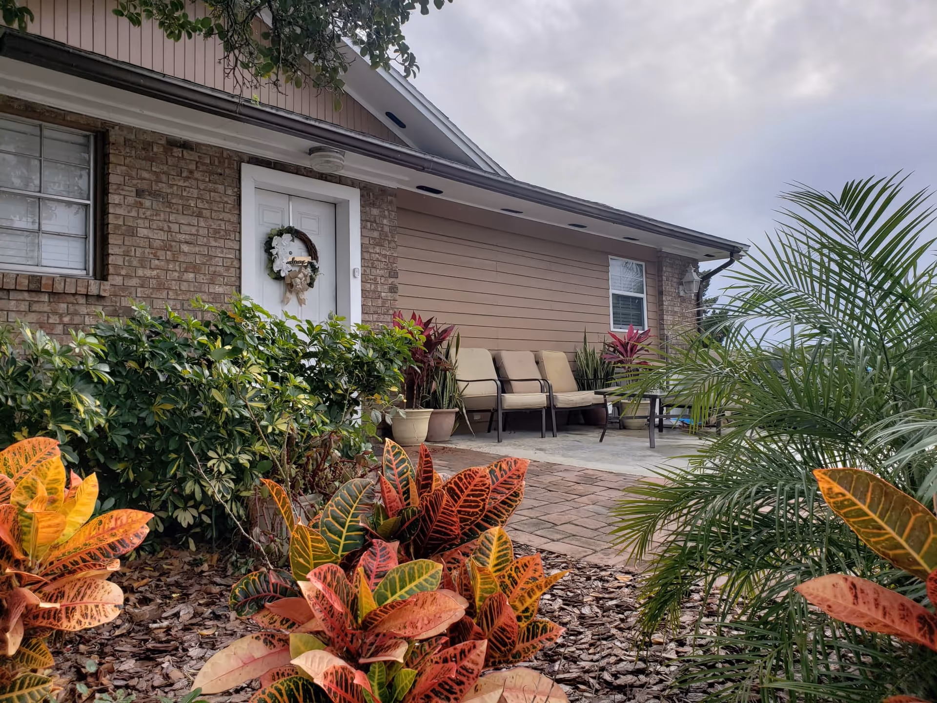 Front entrance and patio of a single-story building with a wreathed door, potted plants, chairs, and colorful foliage in the foreground.
