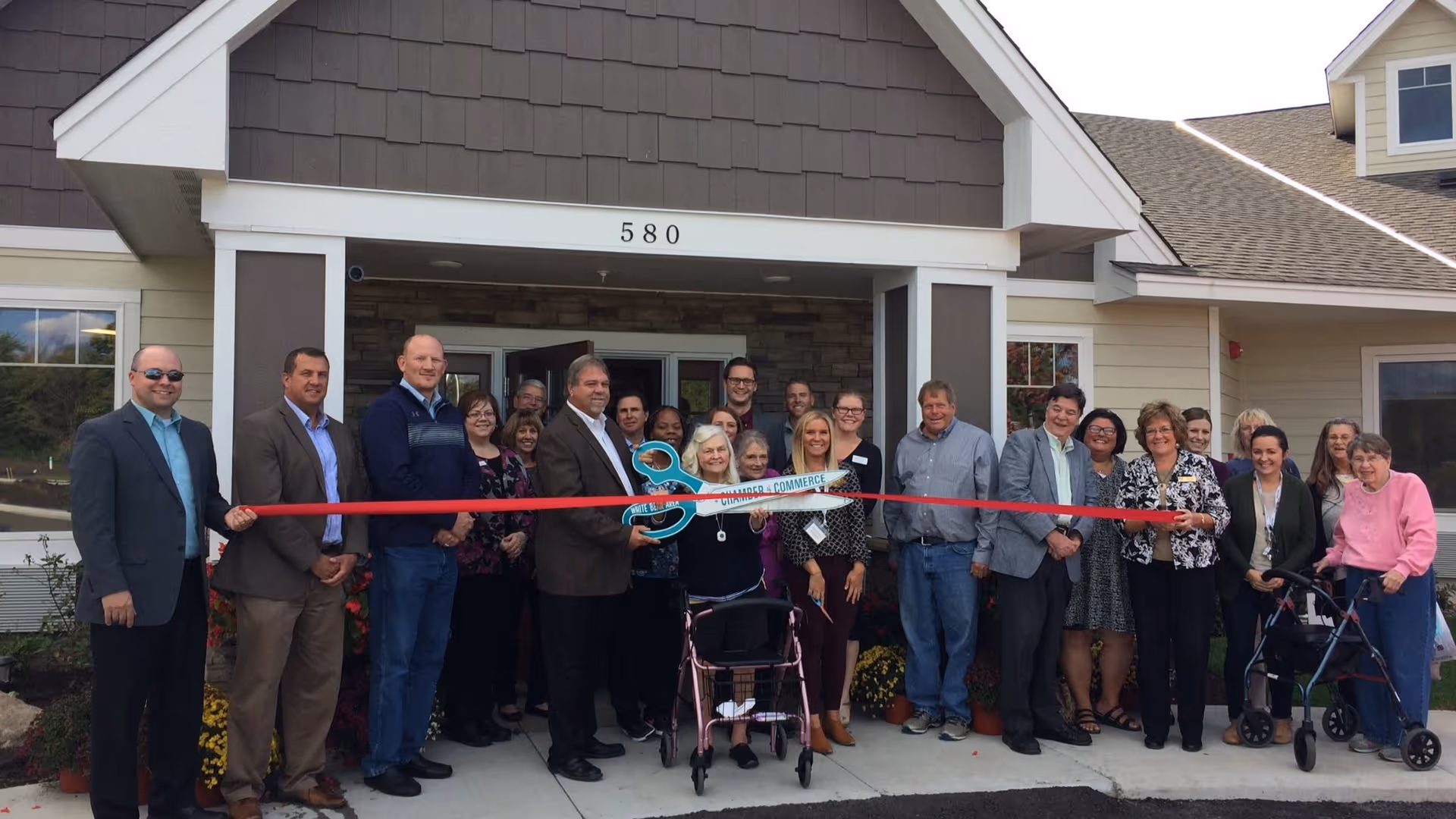 Group of people holding a large red ribbon and oversized scissors for a ribbon-cutting in front of a building entrance numbered 580.