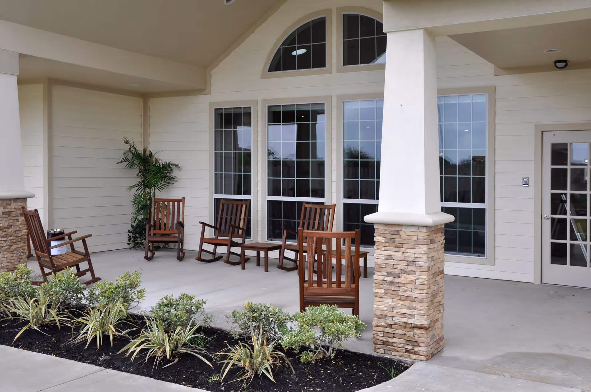 Covered outdoor patio area with several wooden rocking chairs and small tables arranged near large windows and a door. There are plants in a garden bed along the edge of the patio and a stone pillar supporting the roof.