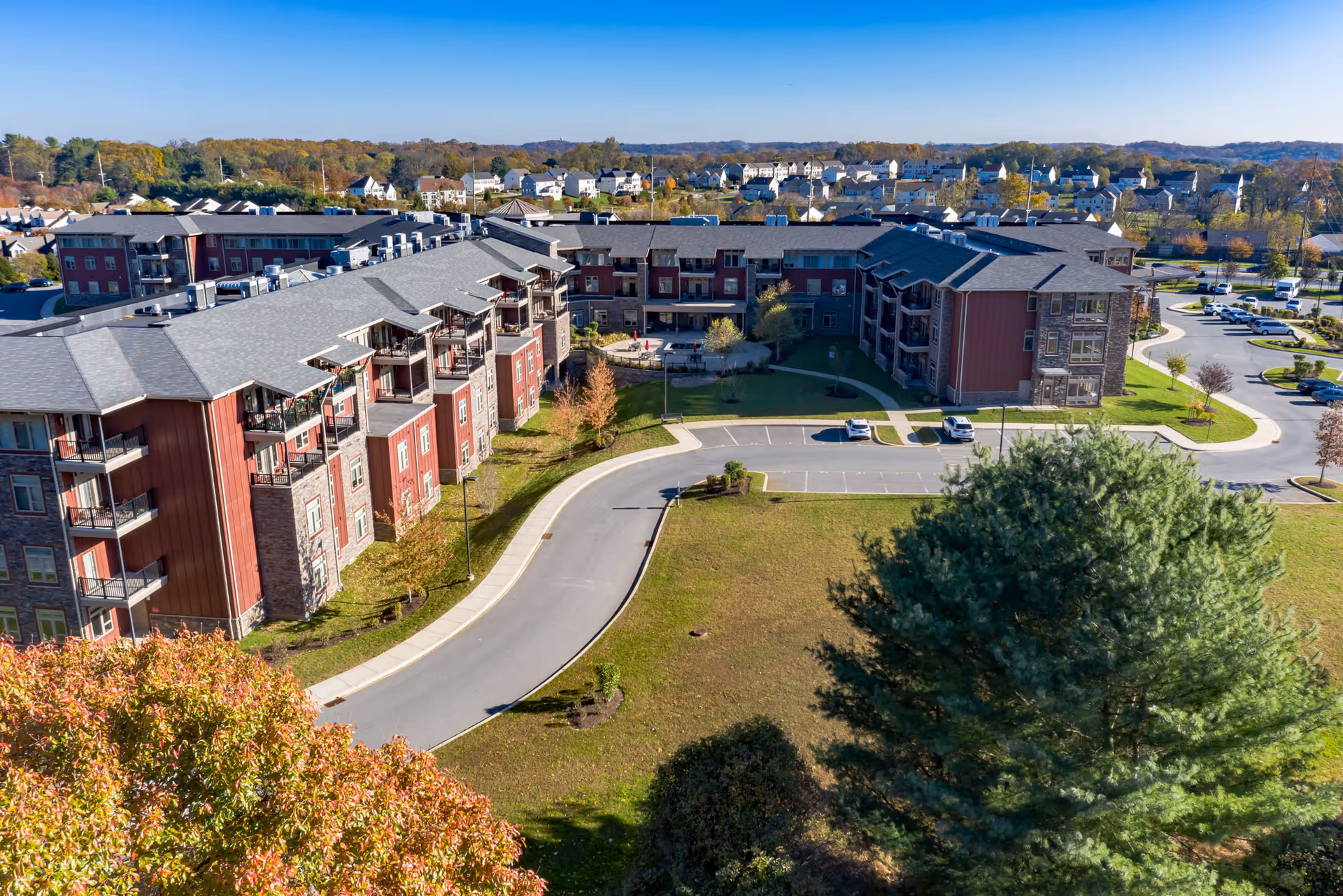 Aerial view of a large senior living facility named The Summit, featuring multiple connected buildings with red and stone exterior walls, surrounded by parking lots, green lawns, and trees with autumn foliage. Residential houses and wooded hills are visible in the background under a clear sky.