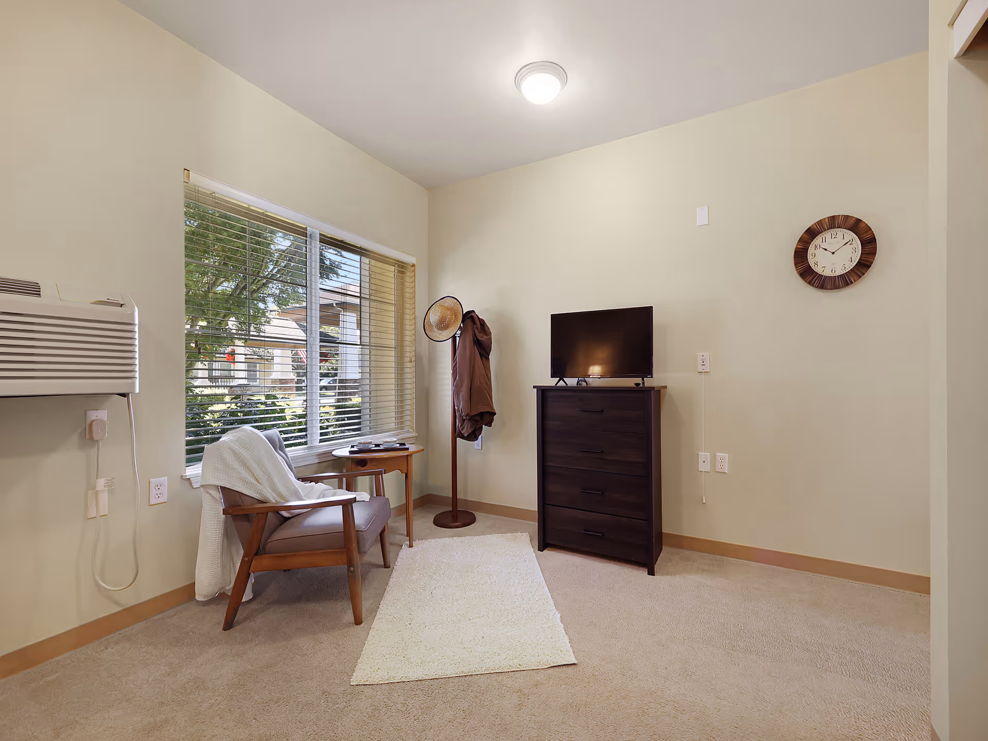 A small, cozy room with beige walls and carpet. There is a wooden armchair with a white blanket draped over it next to a window with blinds. A small wooden side table is beside the chair. Across from the chair is a dark wooden dresser with a flat-screen TV on top. A coat rack with a hat and a jacket stands next to the dresser. A round wall clock hangs on the wall above the dresser. A white rug lies on the floor in front of the dresser and chair. An air conditioning unit is mounted on the wall near the window.