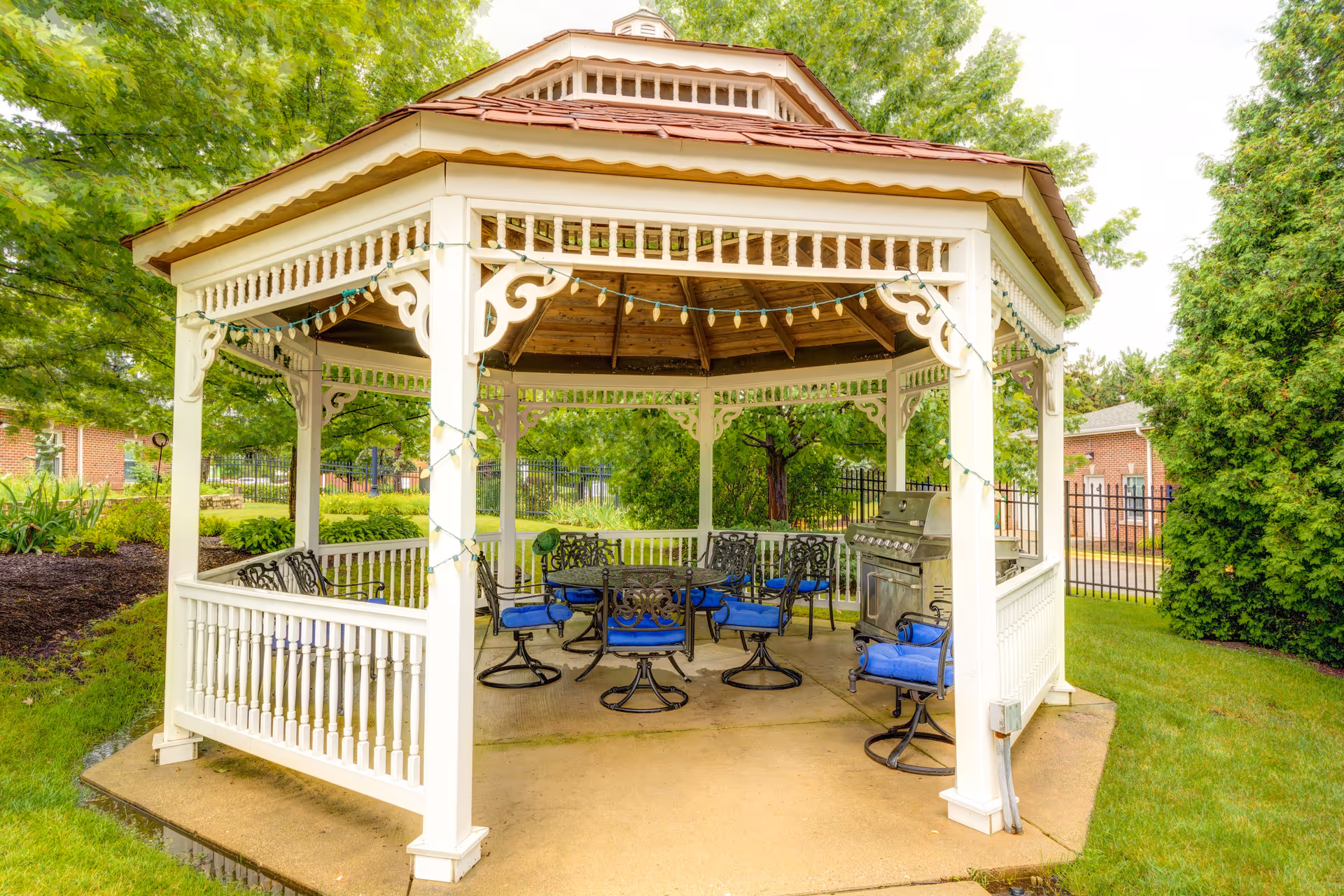 A white wooden gazebo with decorative trim and string lights, containing a round metal table surrounded by six metal chairs with blue cushions, and a stainless steel grill on a concrete patio, set in a green garden area with trees and grass.