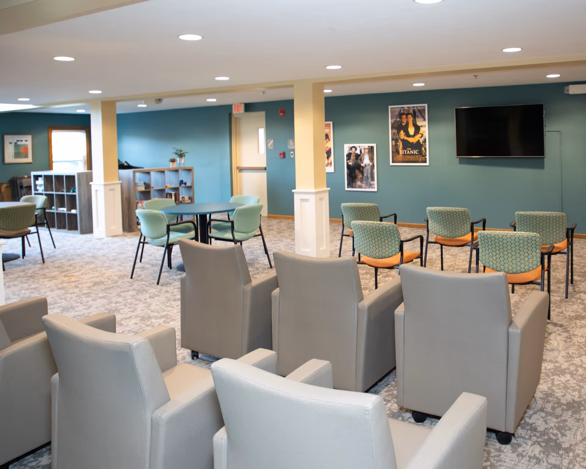 A senior living facility common area with multiple seating arrangements including gray armchairs and green patterned chairs. There is a round table with chairs, a wall-mounted TV, and movie posters on a teal wall. The room has carpeted flooring and recessed ceiling lights.