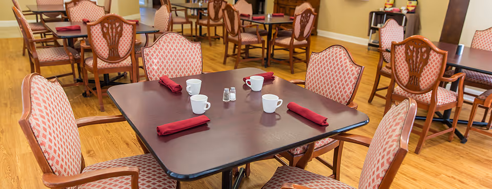 Wood-floored dining room with square tables set with cups, salt and pepper shakers, and red napkins surrounded by patterned upholstered chairs.