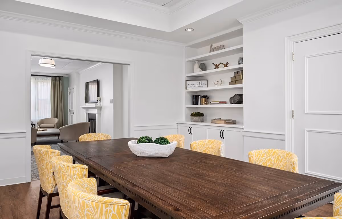 A bright dining room with a large dark wooden table surrounded by yellow patterned chairs. In the background, there is a white built-in shelving unit with decorative items and a doorway leading to a living room area with beige chairs and a fireplace with a mounted TV above it.