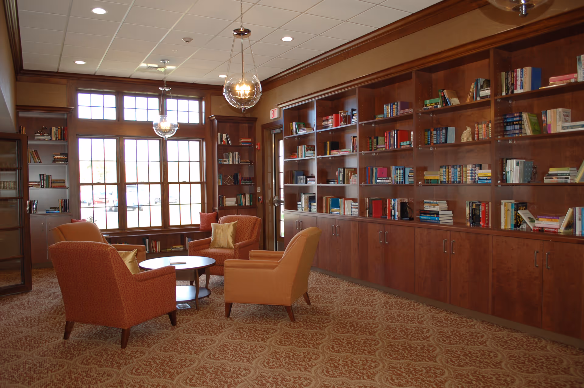 A cozy library or reading room with large wooden bookshelves filled with books along one wall, three orange upholstered armchairs arranged around a small round wooden table, large windows letting in natural light, and two hanging globe light fixtures from the ceiling.