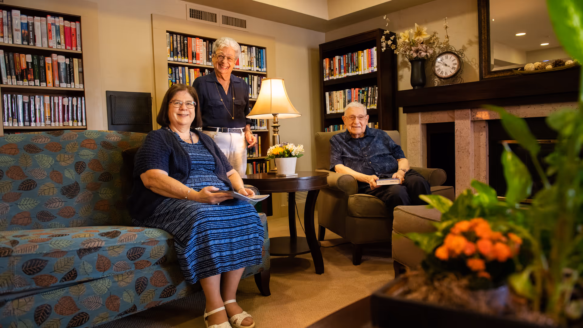 Three elderly people in a cozy living room with bookshelves filled with books. One woman is sitting on a patterned couch holding a book, another woman is standing behind a side table with a lamp and flowers, and an elderly man is sitting in an armchair holding a book. There is a fireplace with a clock and decorative items on the mantel, and a plant with orange flowers in the foreground.