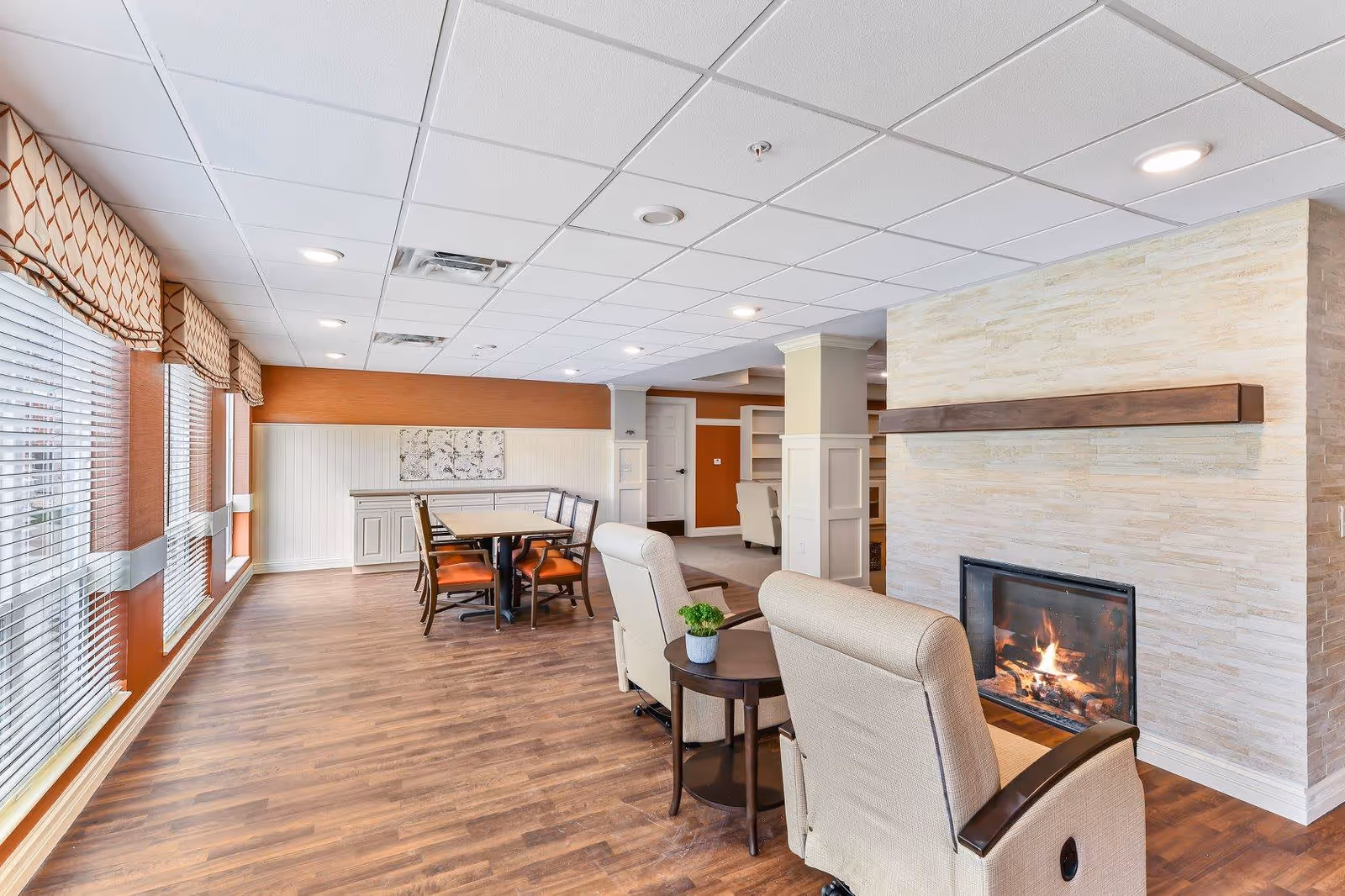 A cozy common area in a senior living facility featuring two beige armchairs with wooden armrests facing a lit fireplace set in a light stone wall. To the left, there is a dining table with six chairs that have orange cushions, positioned near large windows with blinds and patterned valances. The room has wood flooring, white paneled walls with an orange accent wall, and a white ceiling with recessed lighting.