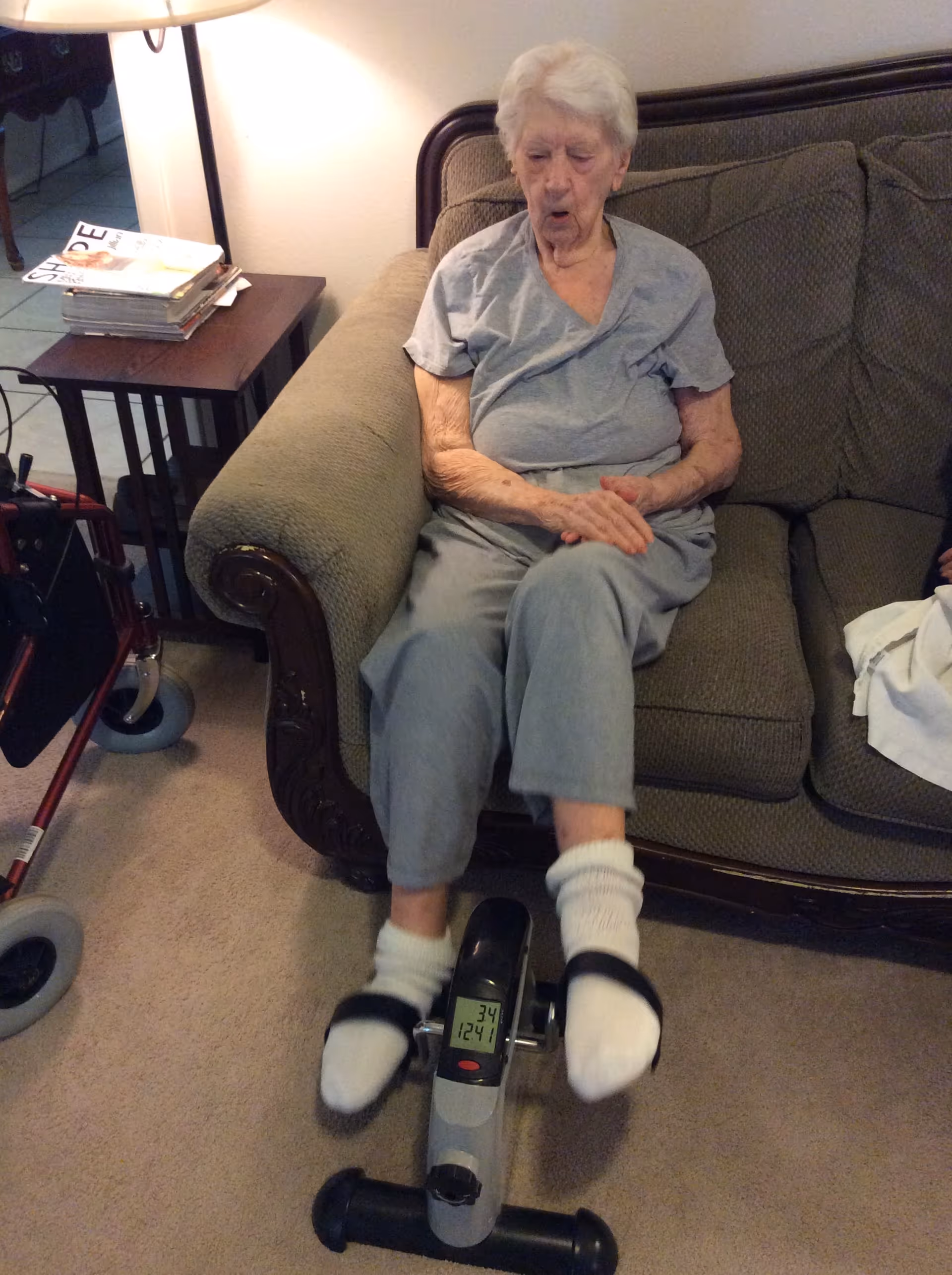 An elderly woman with white hair wearing a gray shirt and pants is sitting on a green upholstered couch, using a small exercise pedal machine with her feet. There is a wooden side table with magazines and a lamp next to the couch, and a red walker is partially visible on the left side.