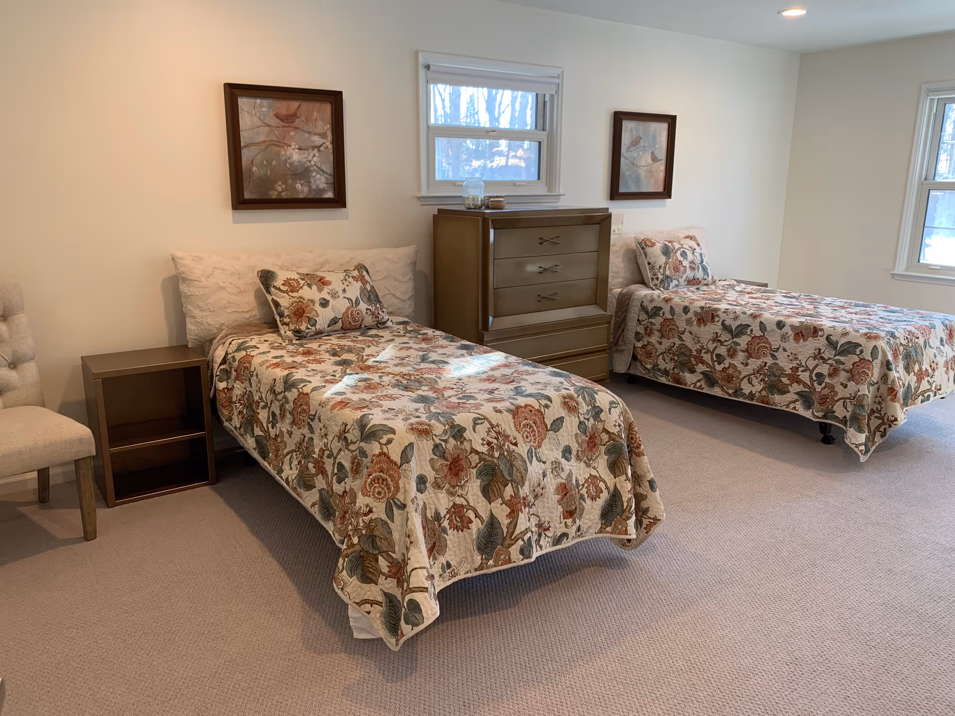 A bedroom with two single beds covered in floral patterned bedspreads. Each bed has a matching pillow and a white headboard. Between the beds is a wooden chest of drawers, and on the left side of the left bed is a small wooden nightstand and a beige upholstered chair. Two framed pictures hang on the wall above the beds, and there are two windows letting in natural light.