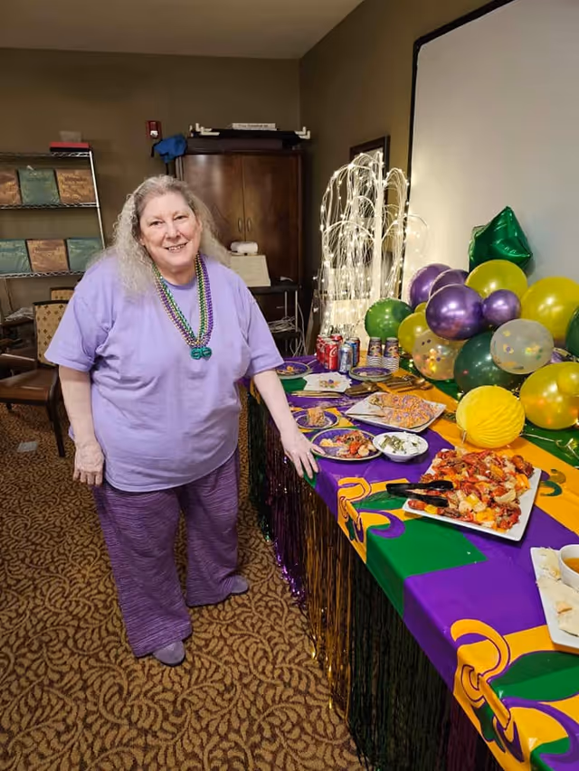 A woman in purple stands beside a decorated buffet table with balloons, plates of food, and drinks in a communal room.