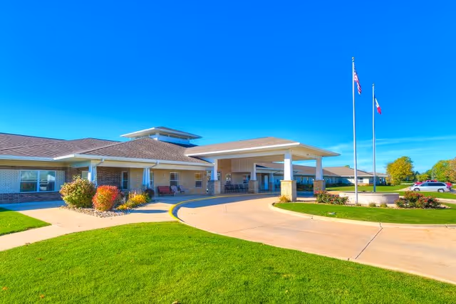 Exterior view of Addington Place of Burlington senior living facility showing a single-story building with a covered entrance, a curved driveway, two flagpoles with flags, well-maintained green lawn, and clear blue sky.