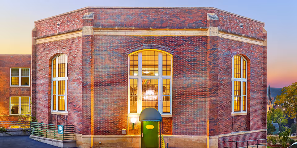 Exterior view of a large brick building with tall arched windows illuminated from inside during sunset. The building has a green door with a small awning above it and a light fixture beside the door. Trees and a distant church steeple are visible in the background.
