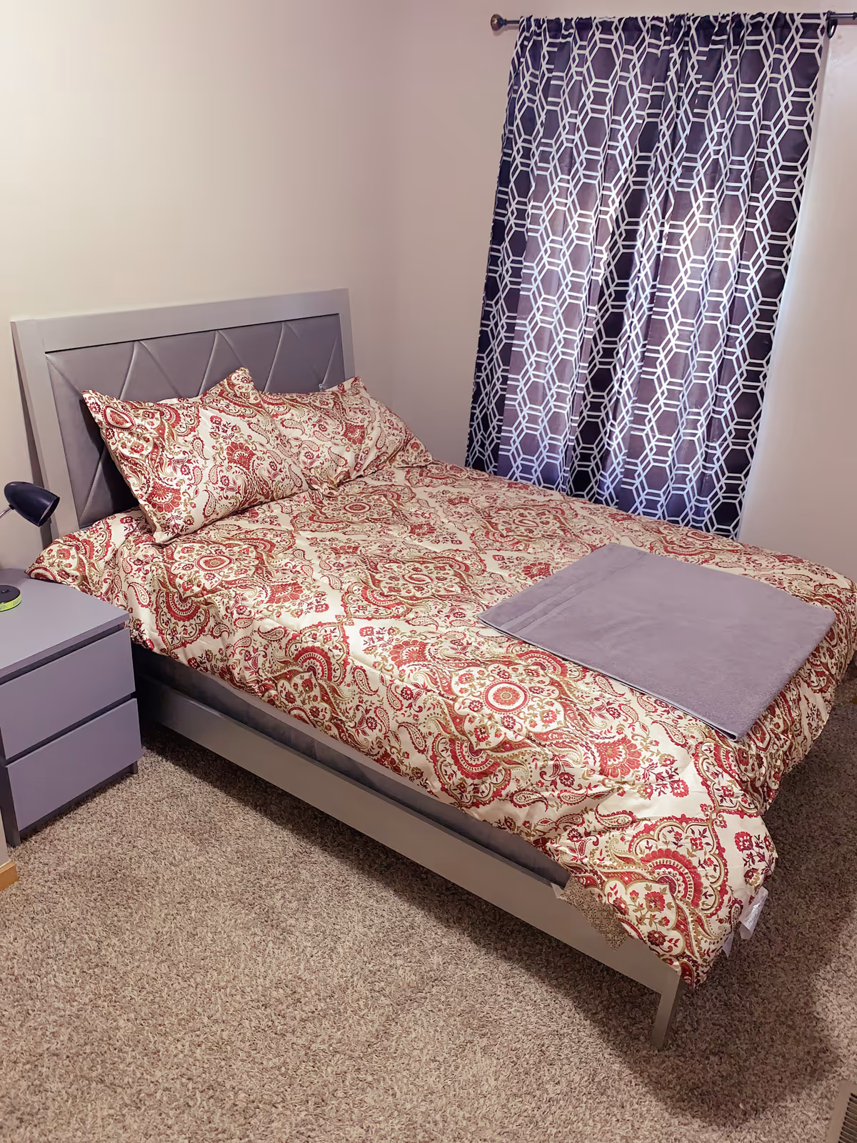 A bedroom with a bed featuring a patterned red and cream bedspread and matching pillows. A folded gray blanket is placed on the bed. Next to the bed is a gray nightstand with a small black lamp. The room has beige carpet and a window with dark curtains featuring a white geometric pattern.