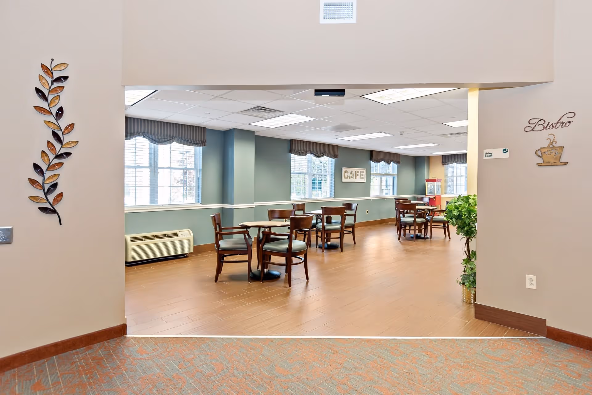 Interior view of a dining area in a senior living facility with several wooden tables and chairs arranged on a wooden floor. The walls are painted green with large windows letting in natural light. Decorative wall art includes a metal leaf sculpture on the left and a sign that says 'Bistro' with a coffee cup on the right. A sign on the far wall reads 'CAFE'.