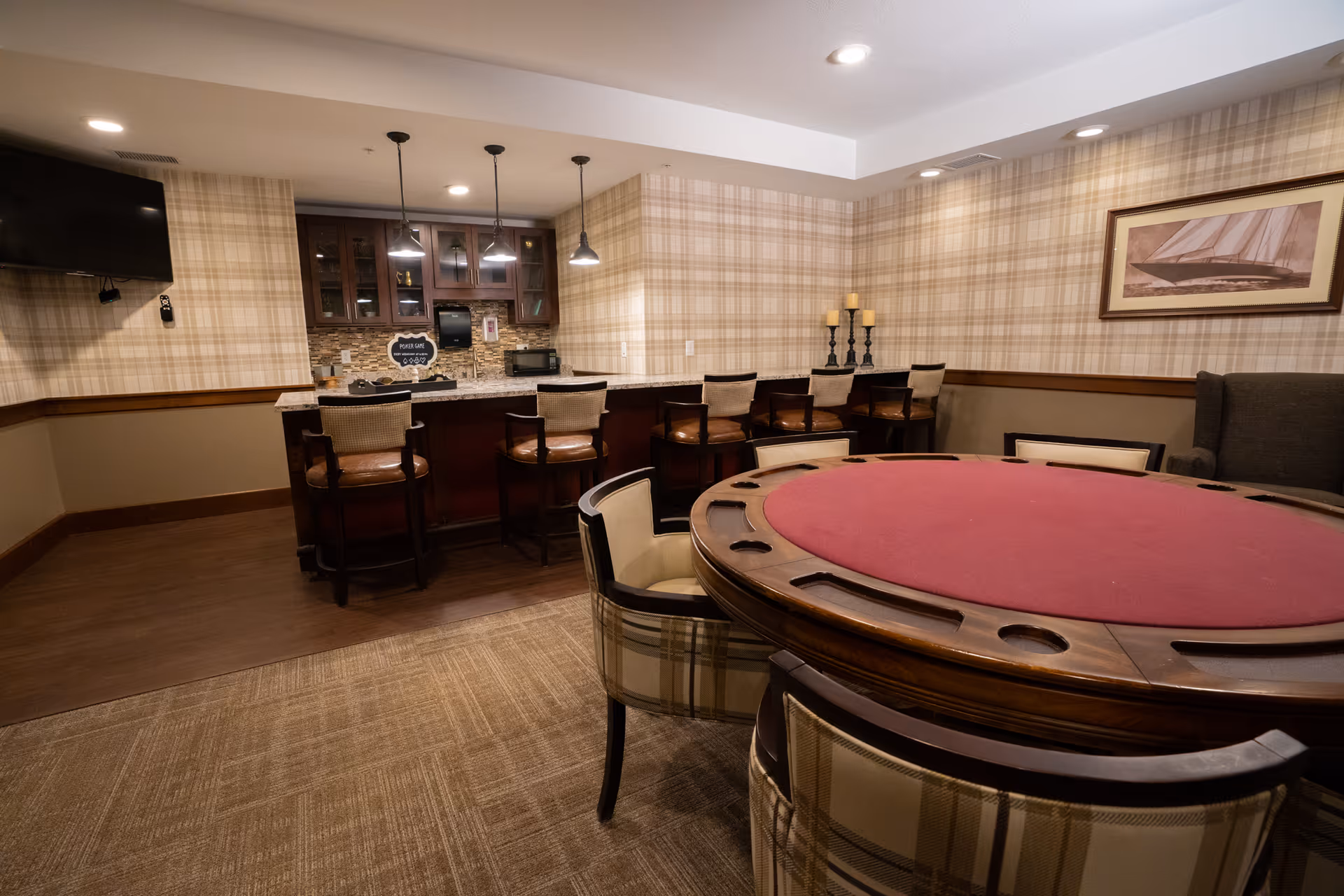 A cozy interior room featuring a round poker table with a red felt top surrounded by cushioned chairs. In the background, there is a bar area with high chairs, pendant lights, and a mounted TV on the wall. The walls have a plaid wallpaper design, and there is a framed picture of a sailboat on the wall.