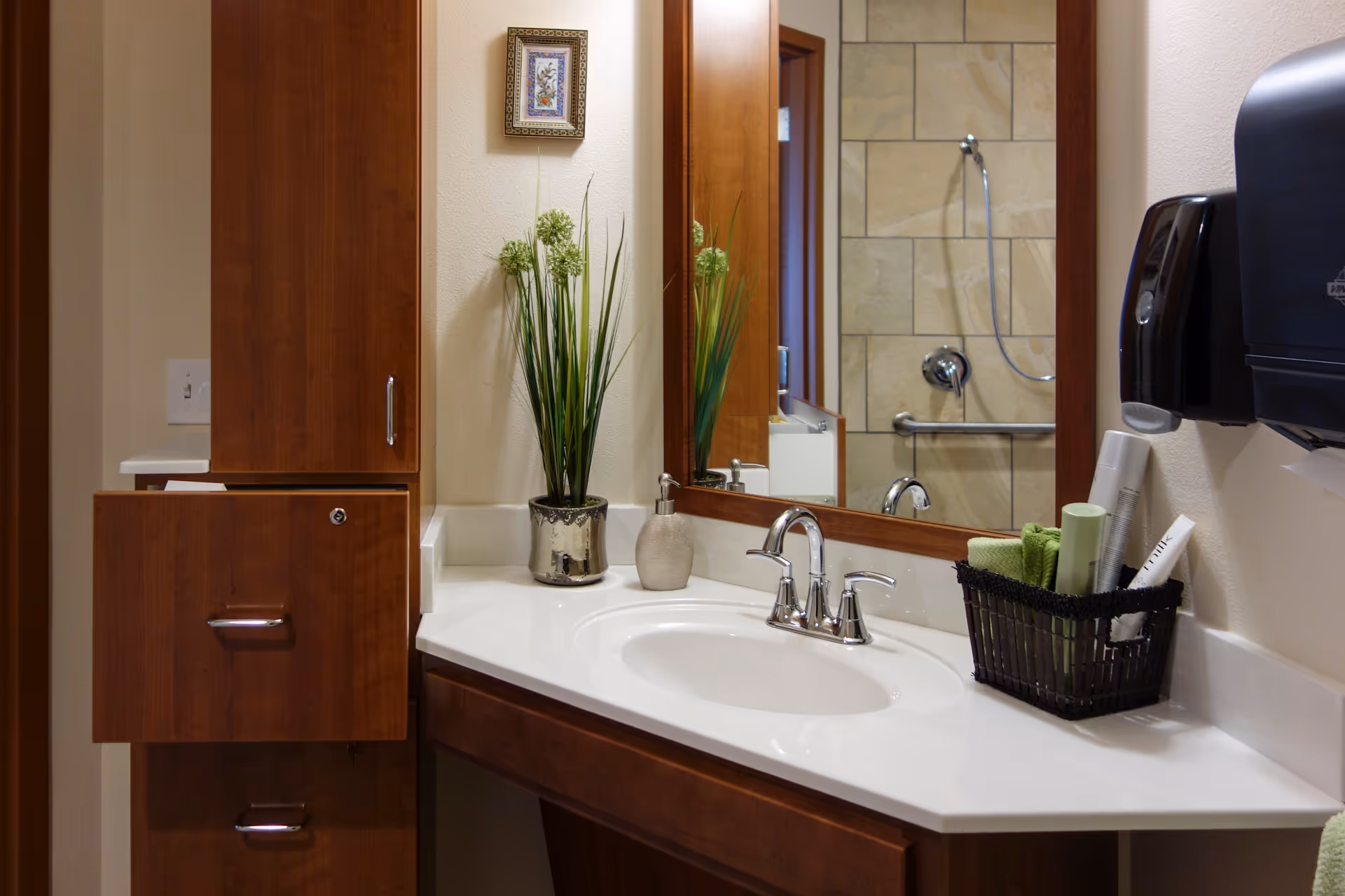 A bathroom vanity with a white countertop and a built-in sink. There is a chrome faucet, a soap dispenser, and a small basket holding toiletries and green towels on the counter. A tall potted plant with green flowers is placed next to the sink. The wall behind the vanity has a large mirror reflecting a tiled shower area with a grab bar and showerhead. Wooden cabinets with silver handles are on the left side of the vanity. A black paper towel dispenser is mounted on the wall to the right.