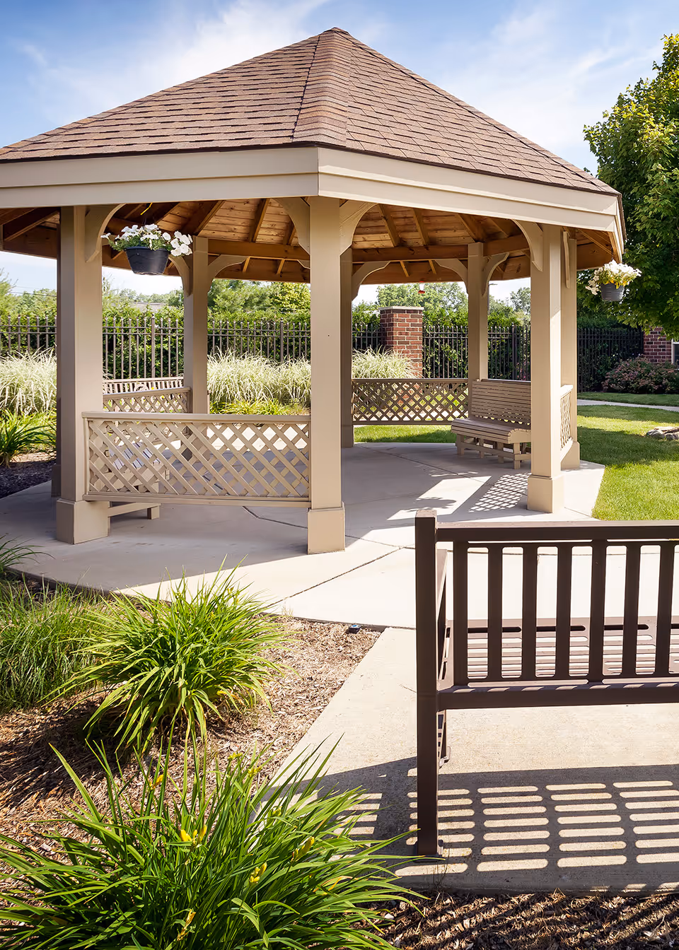 Outdoor gazebo with a shingled roof and beige wooden supports, surrounded by green plants and grass. There are benches inside and outside the gazebo, and hanging flower pots are attached to the structure. The area is sunny with a clear blue sky.