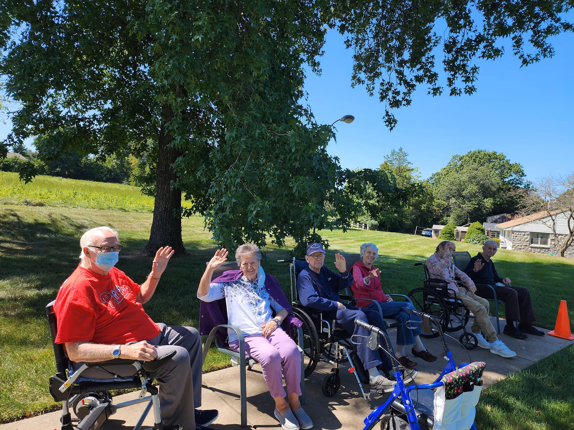 Six elderly individuals sitting outdoors on a sunny day, some in wheelchairs and others in chairs, waving and smiling. They are seated on a sidewalk with green grass and trees in the background under a clear blue sky.