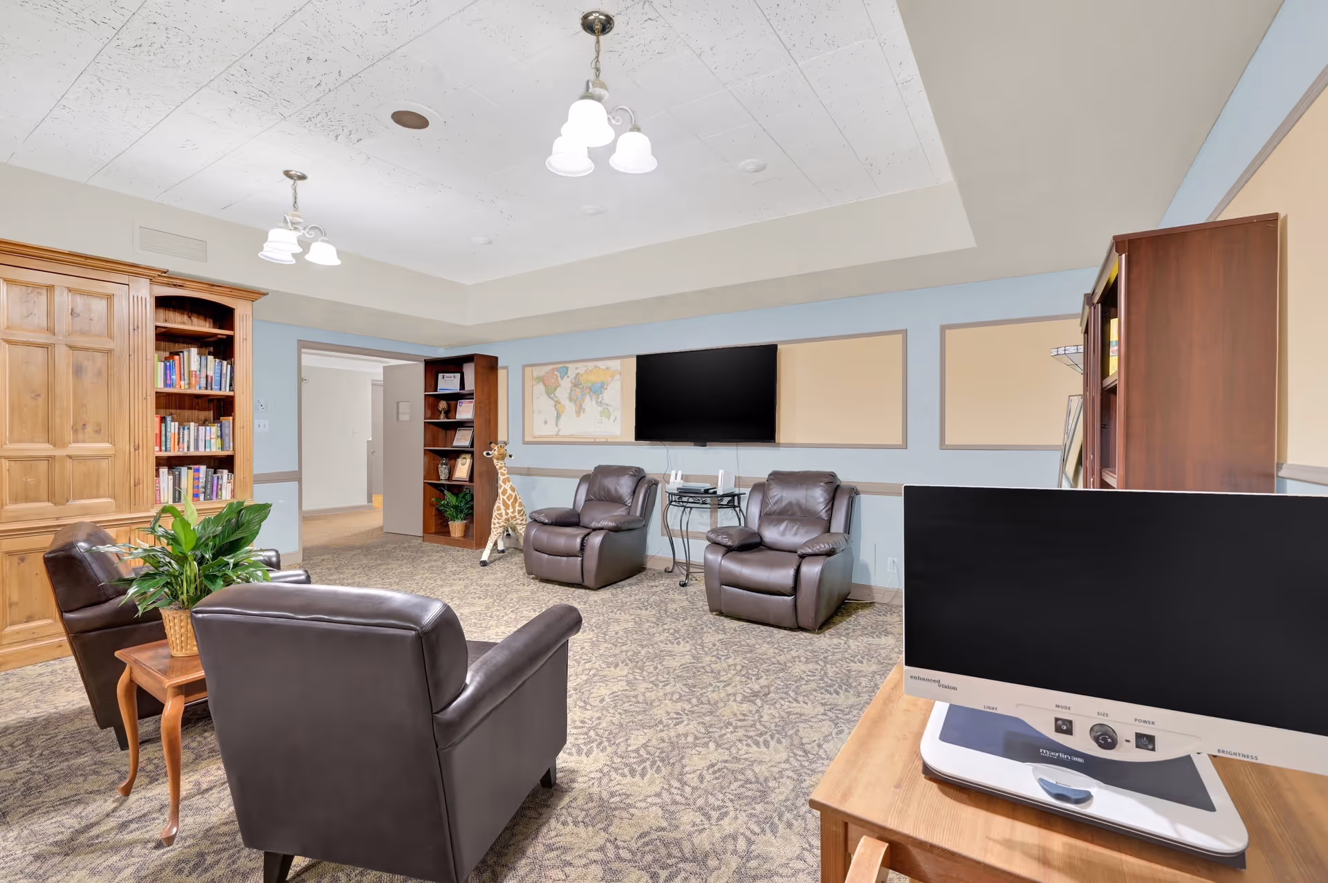 A cozy common area in a senior living facility featuring four dark leather armchairs arranged around a small wooden table with a potted plant. The room has light blue walls with beige bulletin boards, a large flat-screen TV mounted on the wall, bookshelves filled with books, and a decorative giraffe figure near the doorway. The carpet has a patterned design, and the ceiling has white light fixtures.