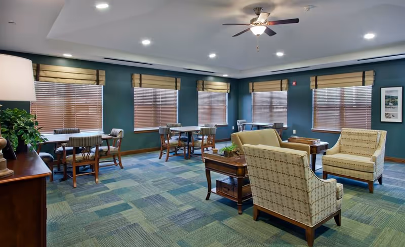 A spacious room with multiple seating areas including tables with chairs and a small lounge area with armchairs and a sofa. The room has green walls, several windows with blinds and valances, a ceiling fan with light, and a patterned carpet in shades of green and blue.