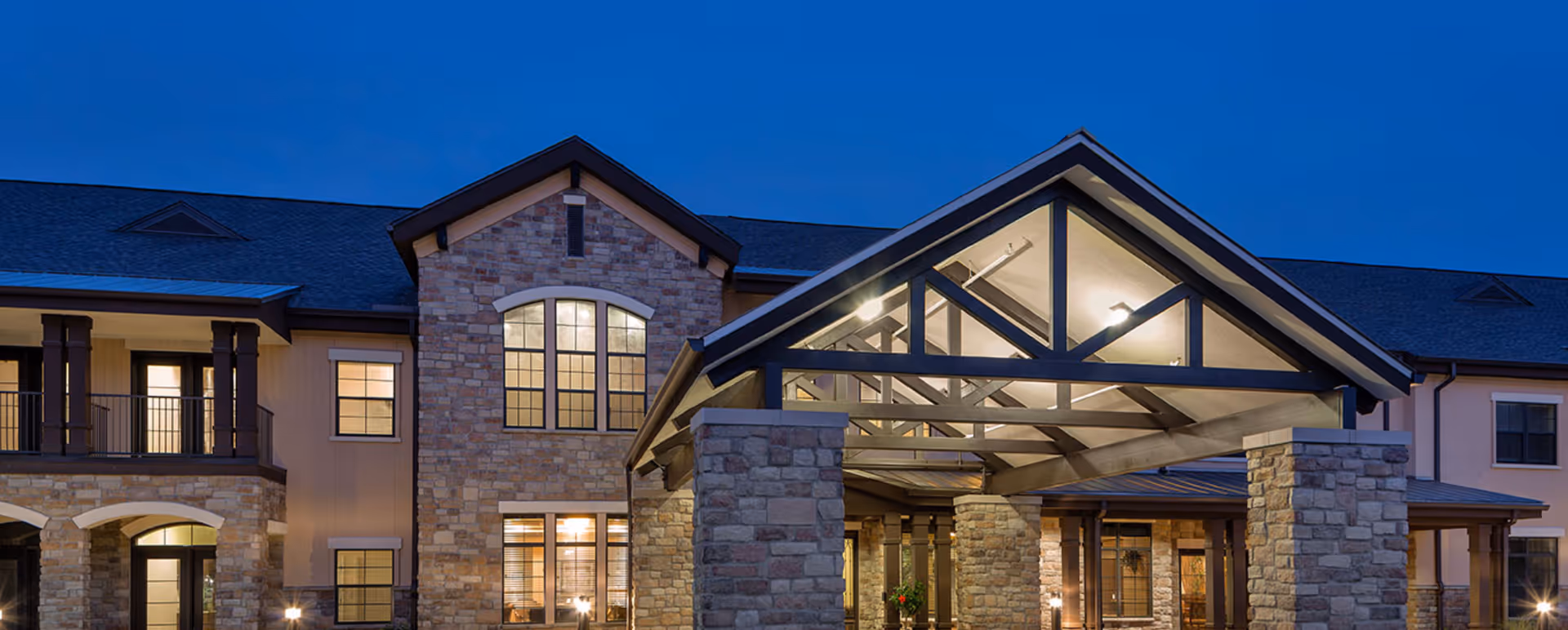 Front exterior of a senior living building at dusk with a stone facade and an illuminated covered entrance.