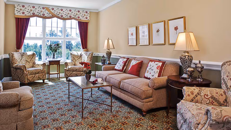 A cozy living room with a large window letting in natural light. The room features a beige sofa with red and floral patterned cushions, two floral upholstered armchairs, a glass coffee table with a small plant and a cup on it, and two side tables with decorative lamps and ornaments. The walls are painted beige with white trim, and there are four framed floral artworks hanging above the sofa. The carpet has a detailed floral pattern.