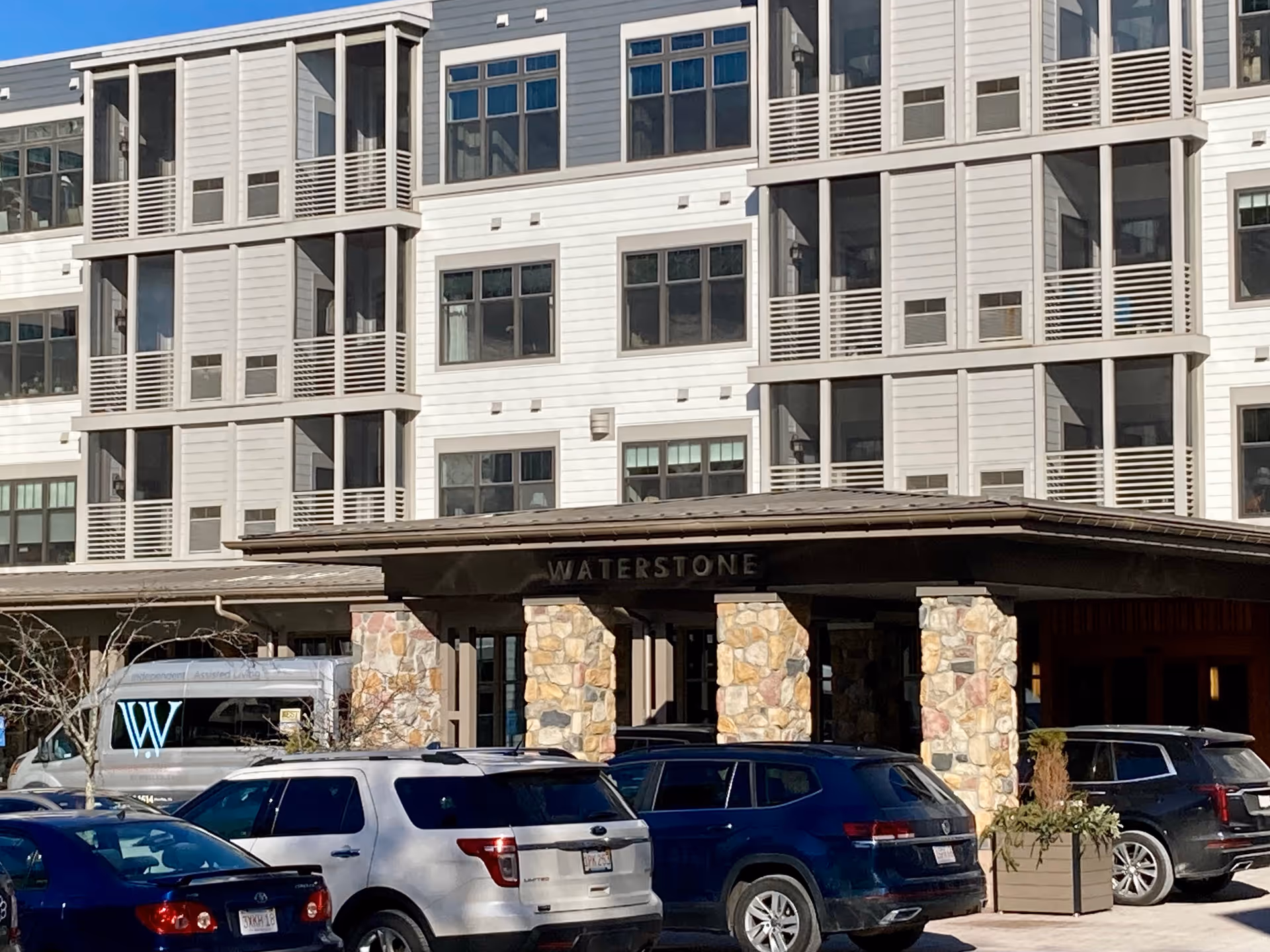 Exterior view of the Waterstone at Wellesley senior living facility showing the entrance with stone pillars and multiple parked cars in front. The building has multiple windows and balconies.