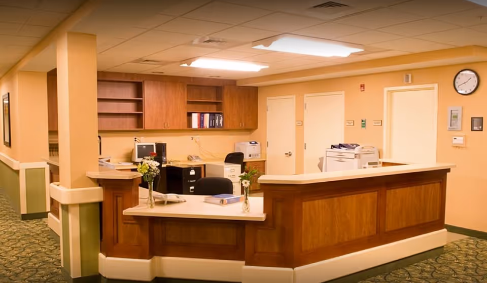 Reception desk area inside a senior living facility with wooden counters, office chairs, computer, printer, and shelves with books. The walls are painted beige with green accents and there is a clock on the wall showing the time as 10:10.