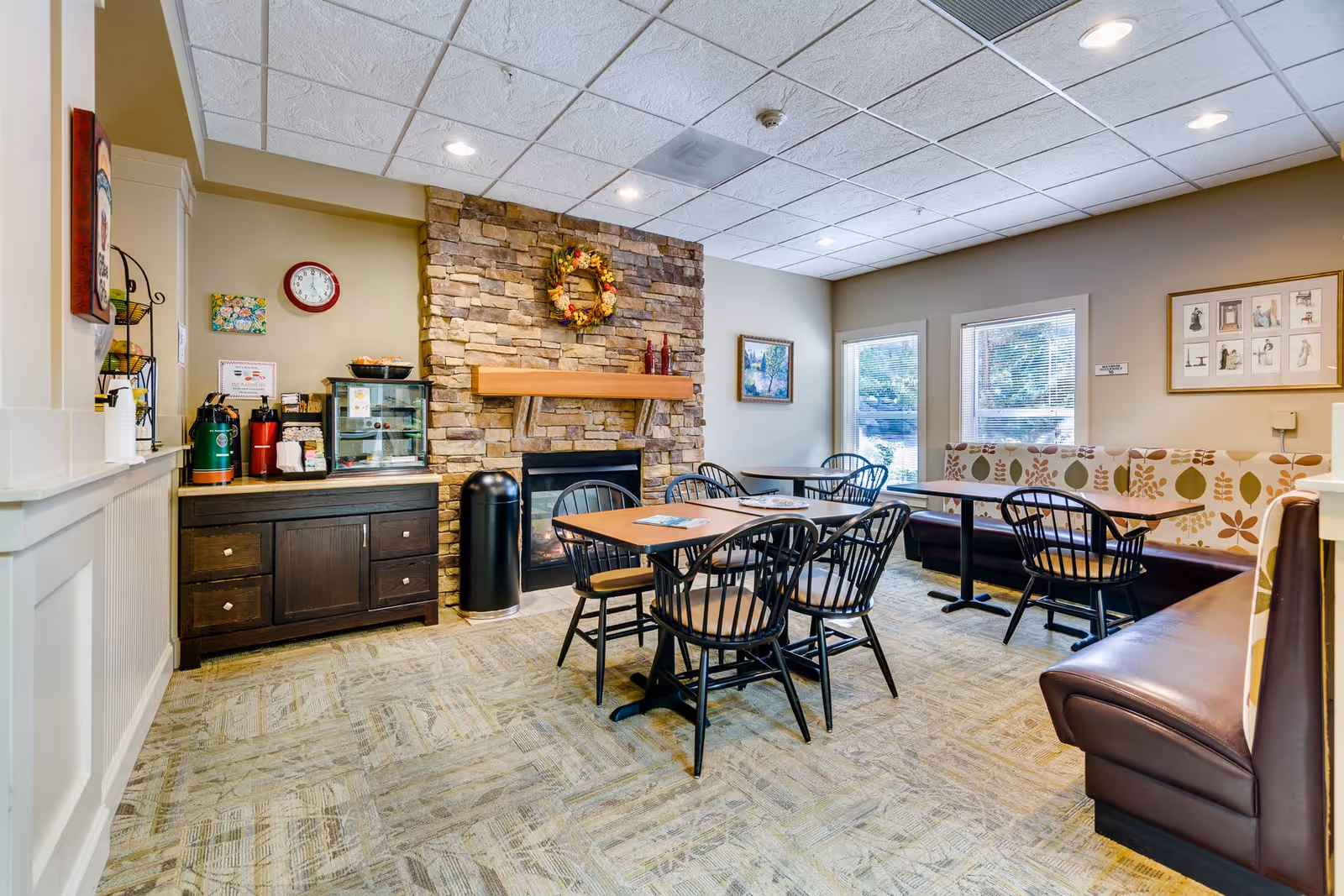 A cozy dining area in Woodland Terrace featuring a stone fireplace with a decorative wreath above it, several tables with black chairs, cushioned bench seating with leaf-patterned upholstery, a cabinet with coffee and tea supplies, and windows letting in natural light.