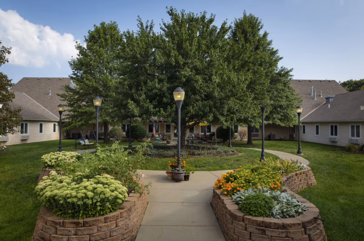 A landscaped courtyard with a large central tree, paved walkways, lamp posts and raised flower beds surrounded by single-story assisted living buildings.