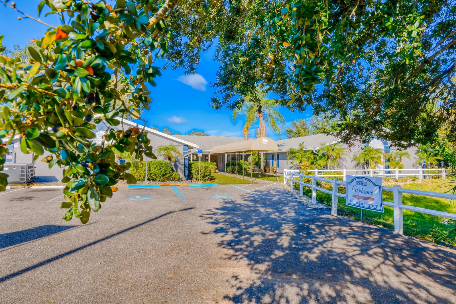 Exterior view of Great American Assisted Living at Tampa showing the main entrance with a covered walkway, surrounded by greenery and trees under a clear blue sky. A white fence lines the driveway leading to the entrance, and a sign near the fence reads 'Welcome Main Entrance'.