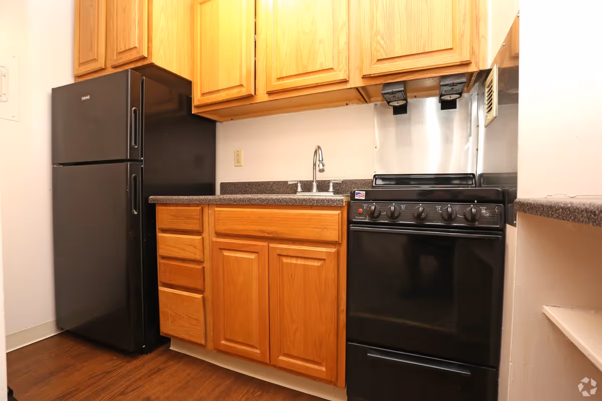 A compact kitchen area featuring wooden cabinets, a black refrigerator, a black stove with control knobs, a stainless steel backsplash behind the stove, a countertop with a sink and faucet, and wooden flooring.