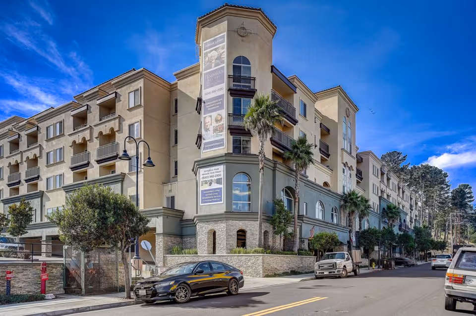 Exterior view of a multi-story senior living facility named Peninsula Del Rey, featuring beige and gray walls, balconies, palm trees, parked cars, and a clear blue sky.