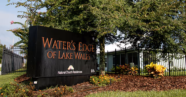 Outdoor view of the entrance sign for Water's Edge of Lake Wales, surrounded by landscaping with flowers, plants, and a tree, with a black metal fence and building in the background.
