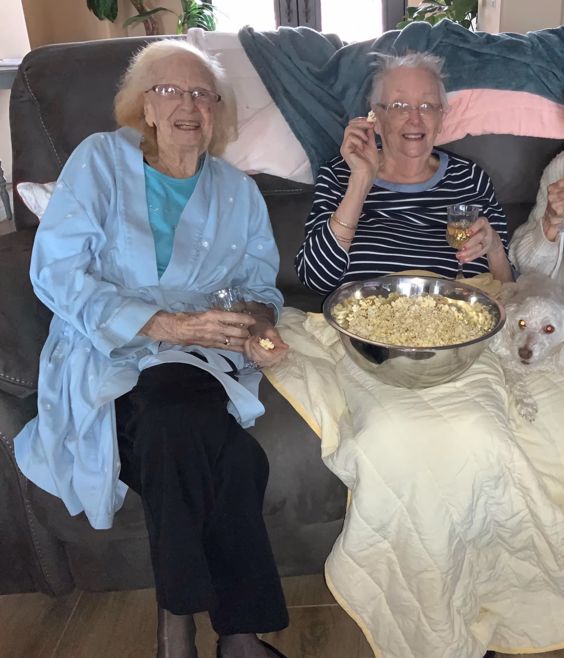 Two elderly women sitting on a couch, smiling and holding glasses of a beverage. One woman is wearing a light blue robe and the other is in a black and white striped shirt. They have a large bowl of popcorn on a blanket on their laps, and a small white dog is lying next to them on the couch.