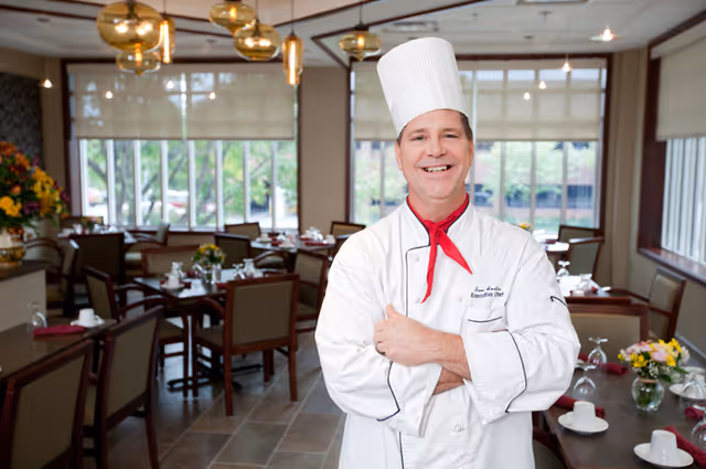 A smiling male chef wearing a white chef's coat and tall white chef hat stands with arms crossed in a well-lit dining room with tables and chairs set for a meal, featuring flowers and tableware.