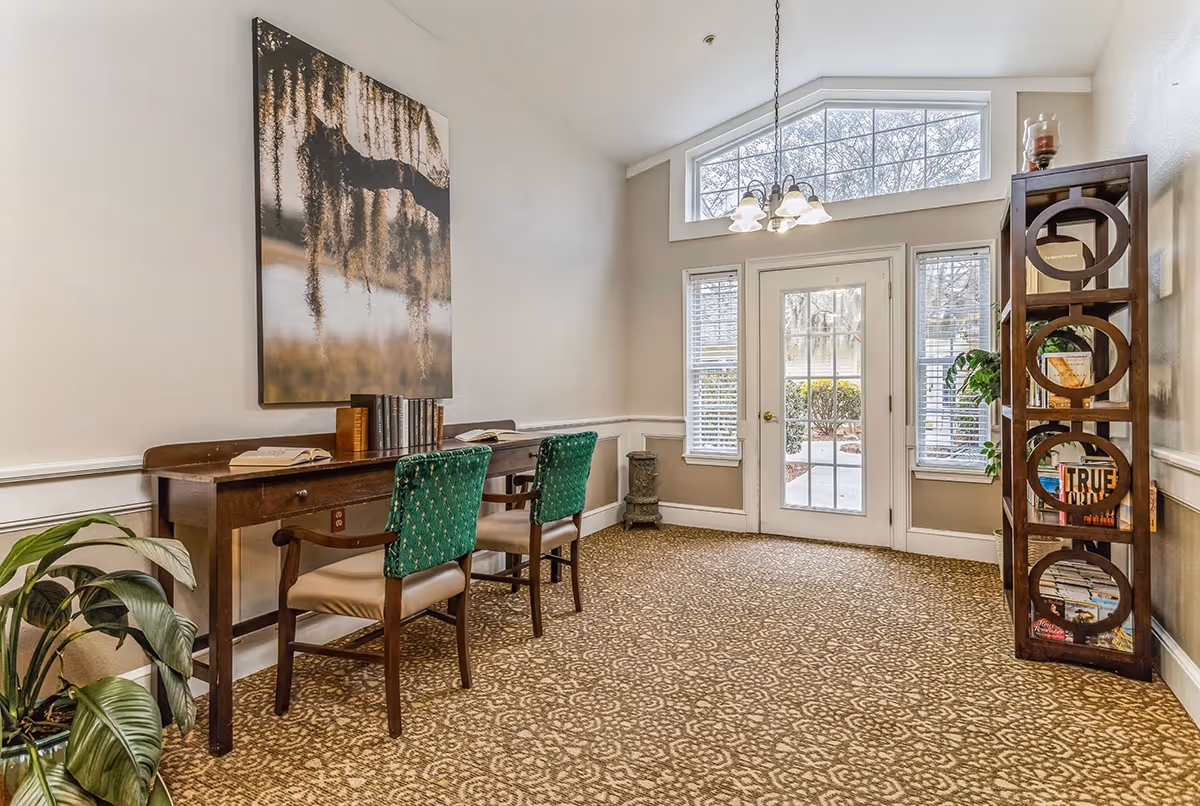 Bright common room with a desk and two green-upholstered chairs, a decorative bookshelf, and a glass door with windows leading outside.