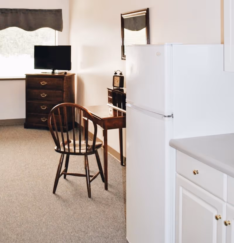 Small studio-style interior showing a kitchenette with a white refrigerator and cabinets, a wooden chair and desk, and a dresser with a TV by a window.