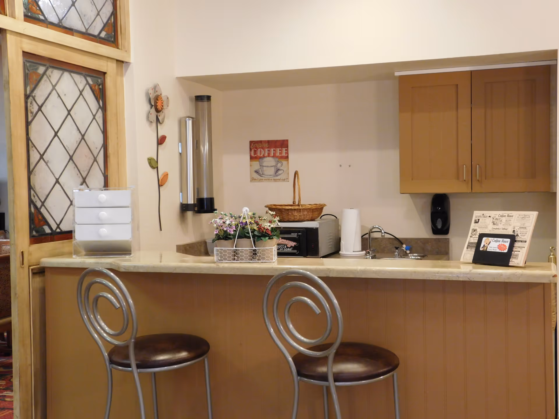 A small kitchen or coffee bar area with a countertop and two metal chairs with circular backrests. The counter has a basket, a microwave, a paper towel holder, a small flower arrangement, and a coffee-themed sign on the wall. There are wooden cabinets above the sink and a decorative stained glass door to the left.