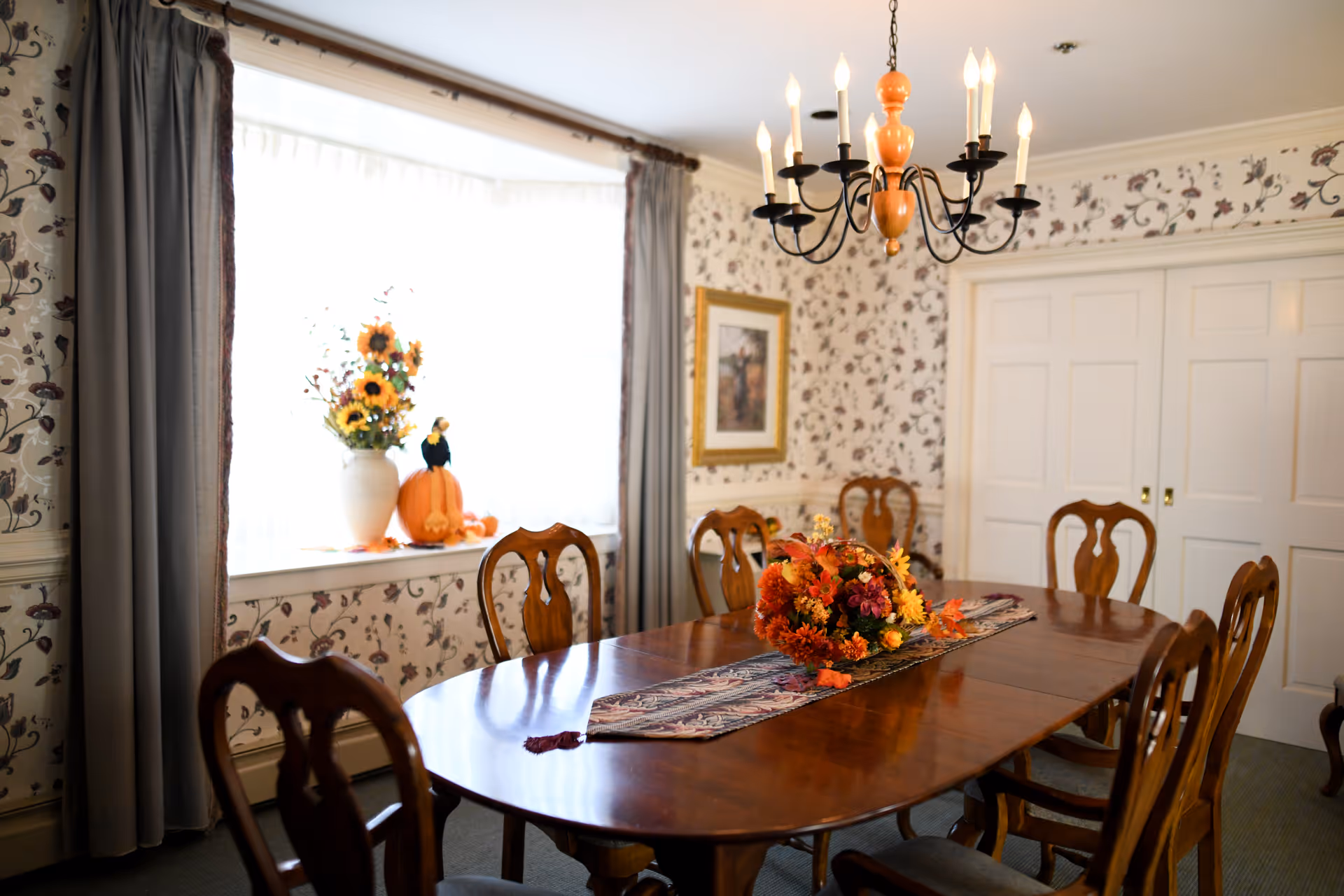 A formal dining room with a wooden table and chairs, a floral centerpiece and chandelier, and a window with fall decorations.