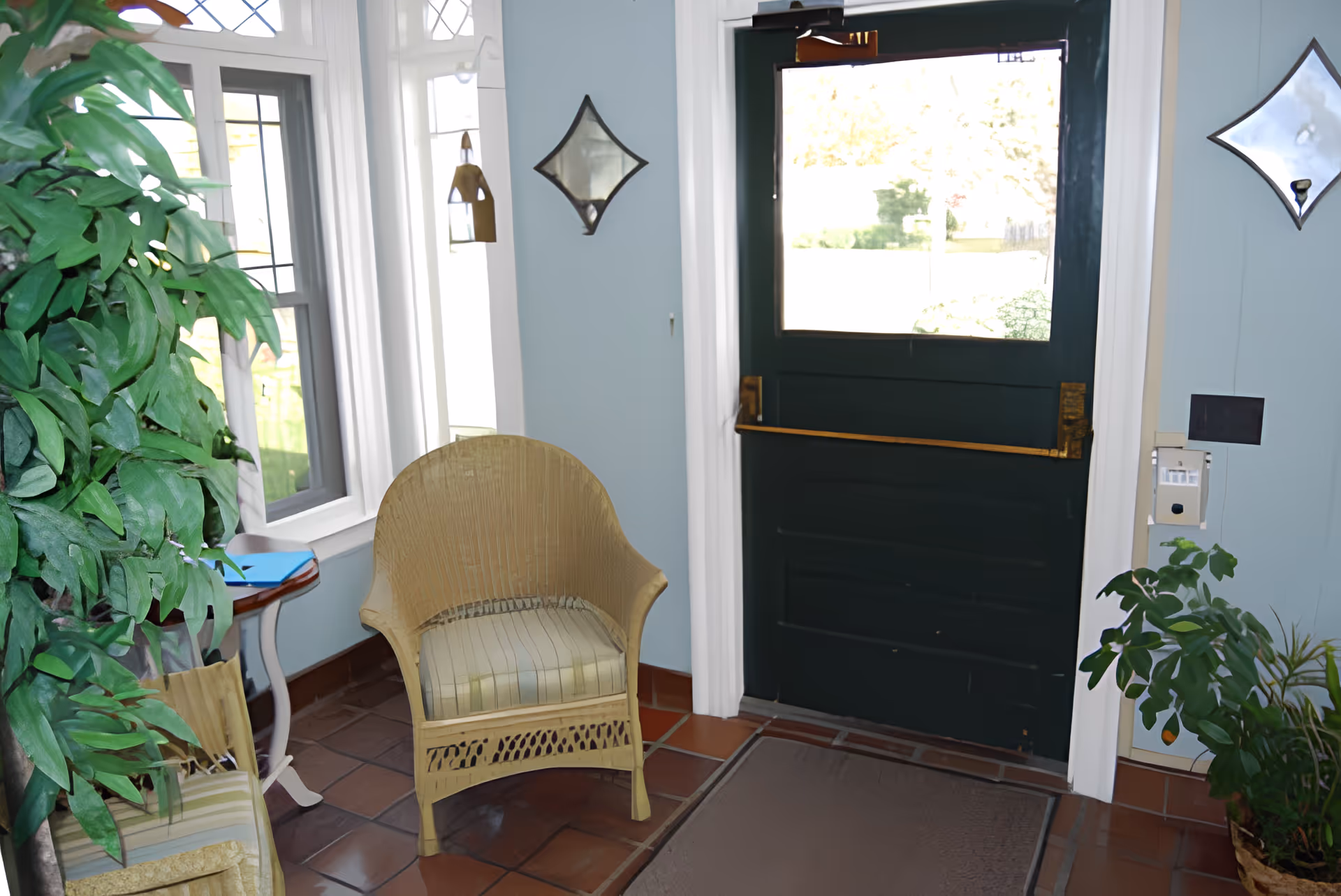 Small entryway with a dark green door featuring a window and a brass push bar. Two wicker chairs with striped cushions are placed near the door, along with a small side table. There are plants on both sides of the door and light blue walls with diamond-shaped mirrors. The floor is tiled with reddish-brown tiles and a brown mat is in front of the door.