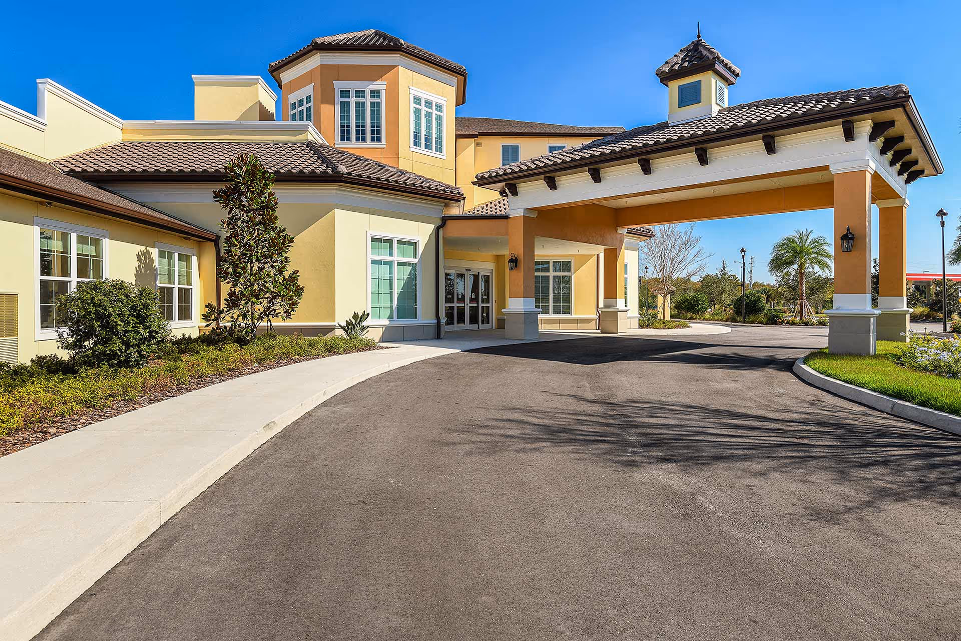Exterior view of a senior living facility building with a covered entrance driveway, beige and yellow walls, tiled roof, and surrounding landscaping including bushes and palm trees under a clear blue sky.