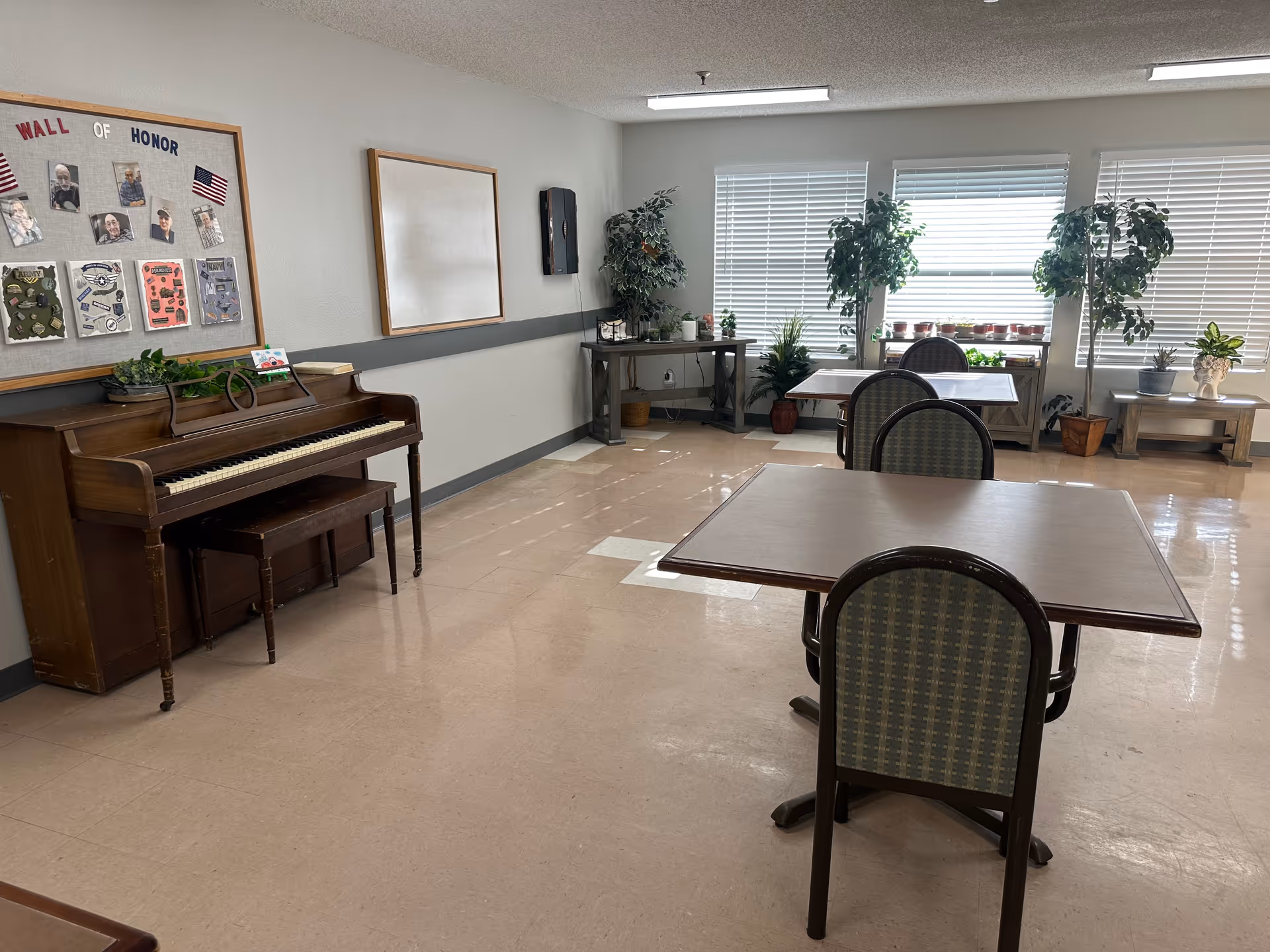 A bright room with several tables and chairs arranged for seating. There is a wooden piano against the left wall beneath a bulletin board titled 'Wall of Honor' displaying photos and small American flags. The room has large windows with blinds, several potted plants on tables and the floor, and a small console table in the corner.