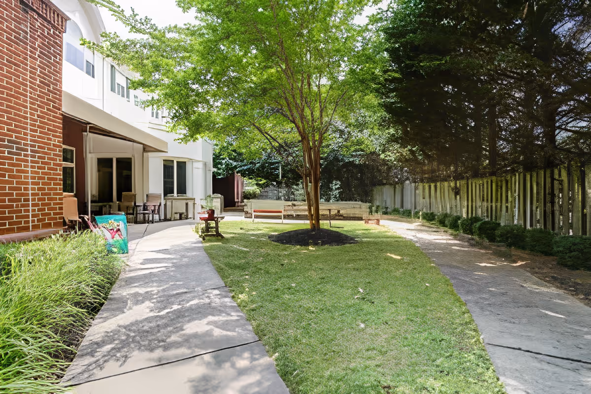 Outdoor garden area at Charter Senior Living of Woodholme Crossing featuring a central tree surrounded by grass, paved walking paths on either side, benches, and a brick and white building on the left side with chairs outside.