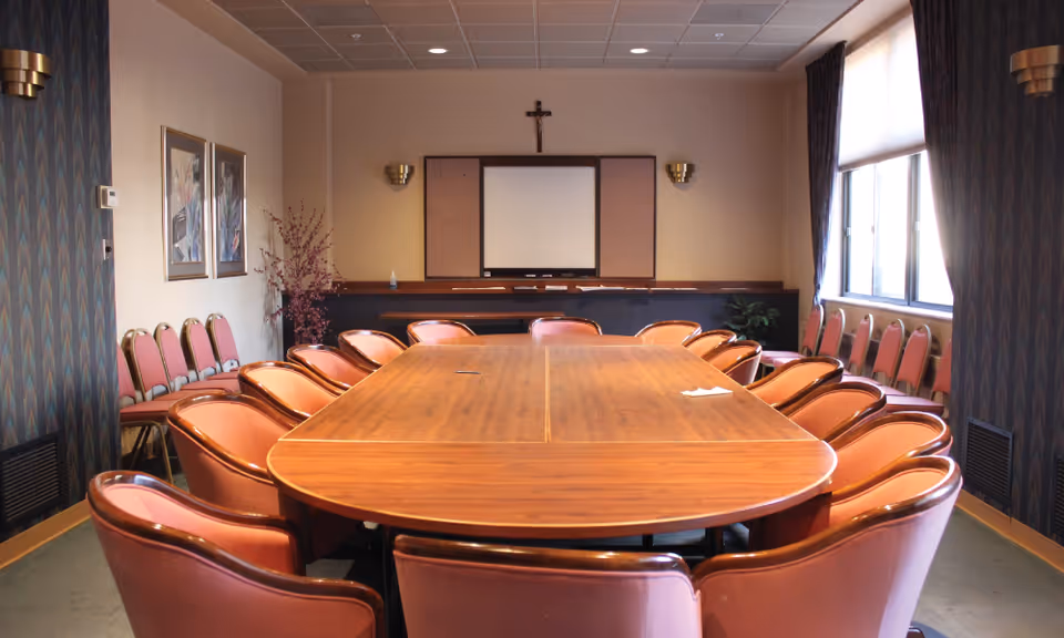 A conference room with a large wooden table surrounded by brown leather chairs. The room has a window with curtains on the right side, framed artwork on the left wall, and a crucifix mounted above a whiteboard at the far end. Additional chairs are lined up against the walls.