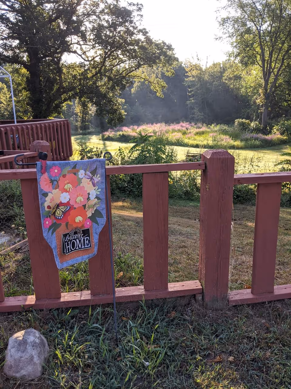 A wooden red fence with a decorative garden flag featuring colorful flowers and a butterfly with the words 'welcome HOME'. Behind the fence is a grassy area with trees and sunlight filtering through, creating a peaceful outdoor scene.