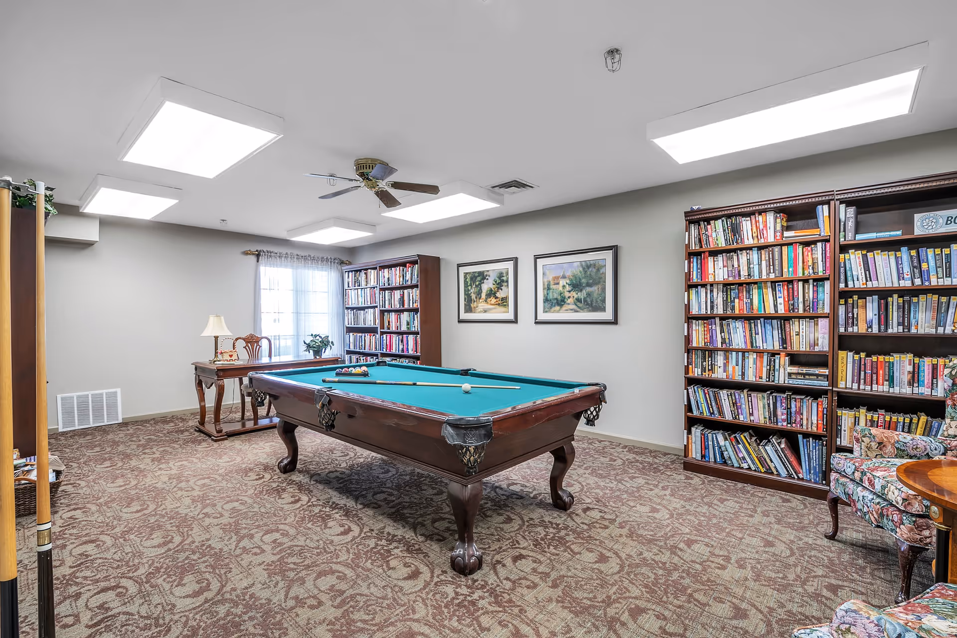 A well-lit recreational room with a pool table in the center, surrounded by bookshelves filled with books. There is a wooden table with a lamp and a chair near a window with sheer curtains. The room has patterned carpet flooring, a ceiling fan, and framed paintings on the wall.