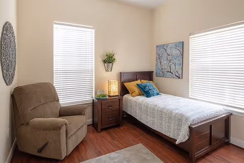 A cozy bedroom with a single wooden bed covered with a light-colored quilt and decorative pillows. Next to the bed is a wooden nightstand with a lamp, a small plant, and a decorative item. There is a comfortable brown recliner chair to the left, two windows with white blinds, a wall-mounted plant, and artwork featuring a tree branch on the wall above the bed. The room has wooden flooring and light-colored walls.
